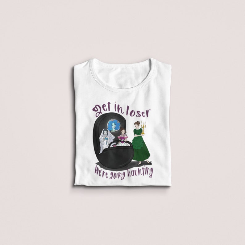 May include: White t-shirt with a graphic featuring a black oval with three figures and the text "Get in loser, we're going haunting" in purple. The figures include a bride, a woman in a green dress, and a woman in a blue dress.