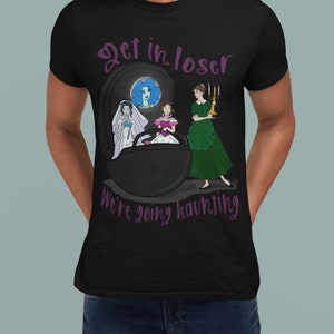 May include: Black t-shirt with a graphic design featuring a tombstone and three cartoon women. The text "Get in loser, we're going haunting" is in purple script. One woman is in a wedding dress, another in a pink dress, and the third in a green dress.