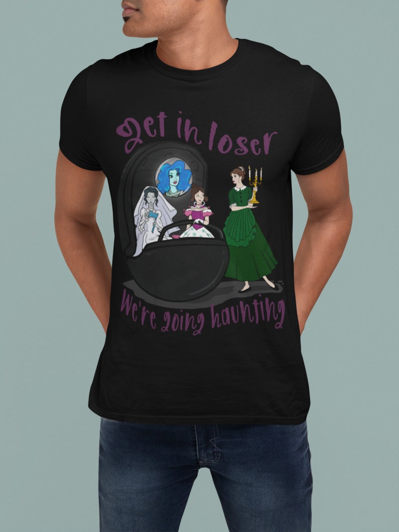 May include: Black t-shirt with a graphic design featuring a tombstone and three cartoon women. The text "Get in loser, we're going haunting" is in purple script. One woman is in a wedding dress, another in a pink dress, and the third in a green dress.