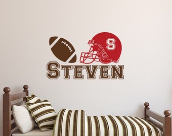 Könnte beinhalten: Wandtattoo mit einem braunen Football, einem roten Helm mit dem Buchstaben "S" und dem Namen "STEVEN" in braunen Blockbuchstaben. Das Tattoo ist für ein Schlafzimmer konzipiert, mit einem Bett und einem Nachttisch.