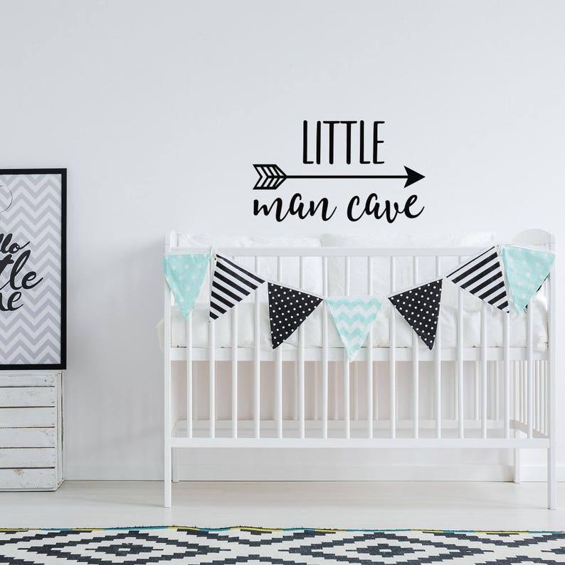 Op de afbeelding: Een witte wieg met een decoratieve slinger van zwart-witte driehoeken en mintgroene chevron vlaggetjes. Op de muur boven de wieg staat de tekst "LITTLE man CAVE" in het zwart. Een ingelijste print en een houten kist zijn ook zichtbaar.