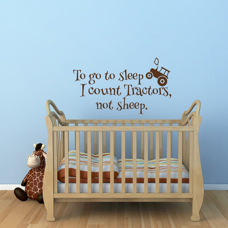 May include: A light brown wooden crib with a striped pillow and a giraffe stuffed animal. The wall above the crib has the text "To go to sleep I count Tractors, not sheep" with a tractor graphic in brown.