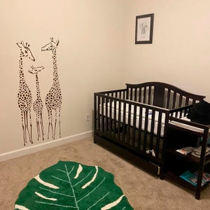 May include: A nursery with a dark brown crib, a green leaf-shaped rug, and a wall decal of three giraffes. A framed picture of an elephant hangs on the wall. The room has a neutral color scheme.