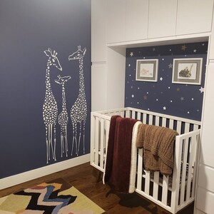 May include: A white crib with a burgundy and brown blanket is set against a navy blue wall with a giraffe stencil. The room features framed artwork and a star-patterned accent wall, creating a nursery theme.