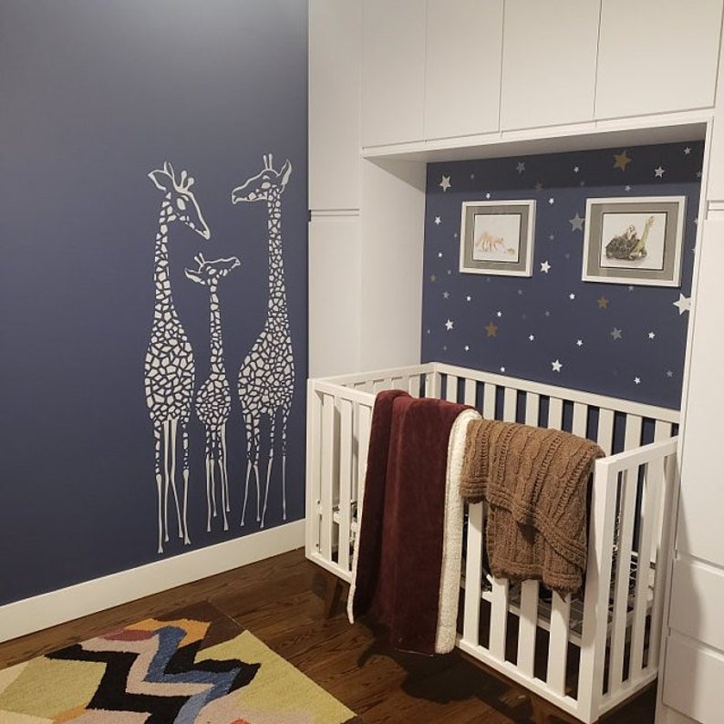 May include: A white crib with a burgundy and brown blanket is set against a navy blue wall with a giraffe stencil. The room features framed artwork and a star-patterned accent wall, creating a nursery theme.