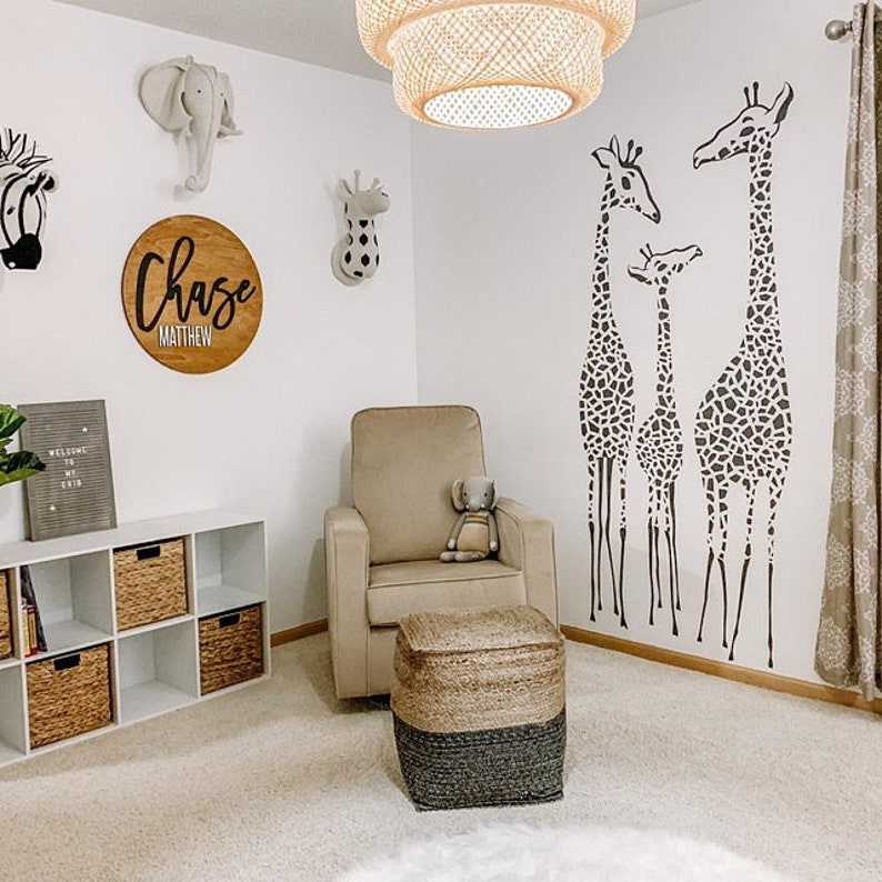 May include: A nursery room with a neutral color scheme. The room features a beige armchair, a woven ottoman, and a white storage unit with wicker baskets. Wall decor includes giraffe decals, an elephant head, and a wooden sign with the name "Chase Matthew".