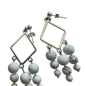May include: A pair of silver dangle earrings with white marble beads. The earrings feature a square shape with a dangling cluster of beads.