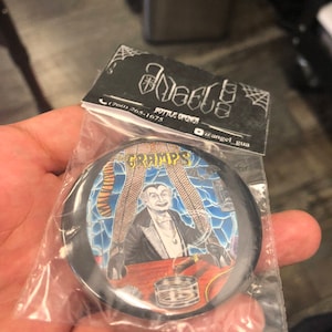 May include: A black and silver bottle opener with a round, colorful image of a vampire with a cigar and two women in fishnet stockings. The image is surrounded by a black border and a spiderweb design. The text "The Gramps" is visible on the image.