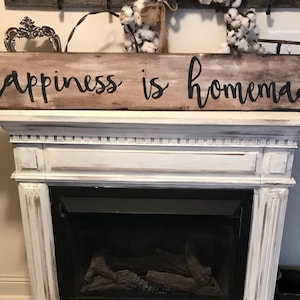 May include: A rustic wooden sign with the text "Happiness is homemade" painted in black. The sign is displayed above a white fireplace mantel.