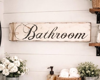 May include: A rectangular wooden sign with a distressed white finish and black cursive text that reads Bathroom. The left side of the sign is wrapped with natural jute twine, creating a decorative accent on the horizontal wall-mounted plaque.