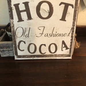 Puede incluir: Un letrero de madera r&uacute;stico con las palabras "HOT Old Fashioned COCOA" en letras marr&oacute;n oscuro. El letrero tiene un fondo blanco y un borde marr&oacute;n oscuro, con un aspecto vintage. Evoca una sensaci&oacute;n de calidez y confort.