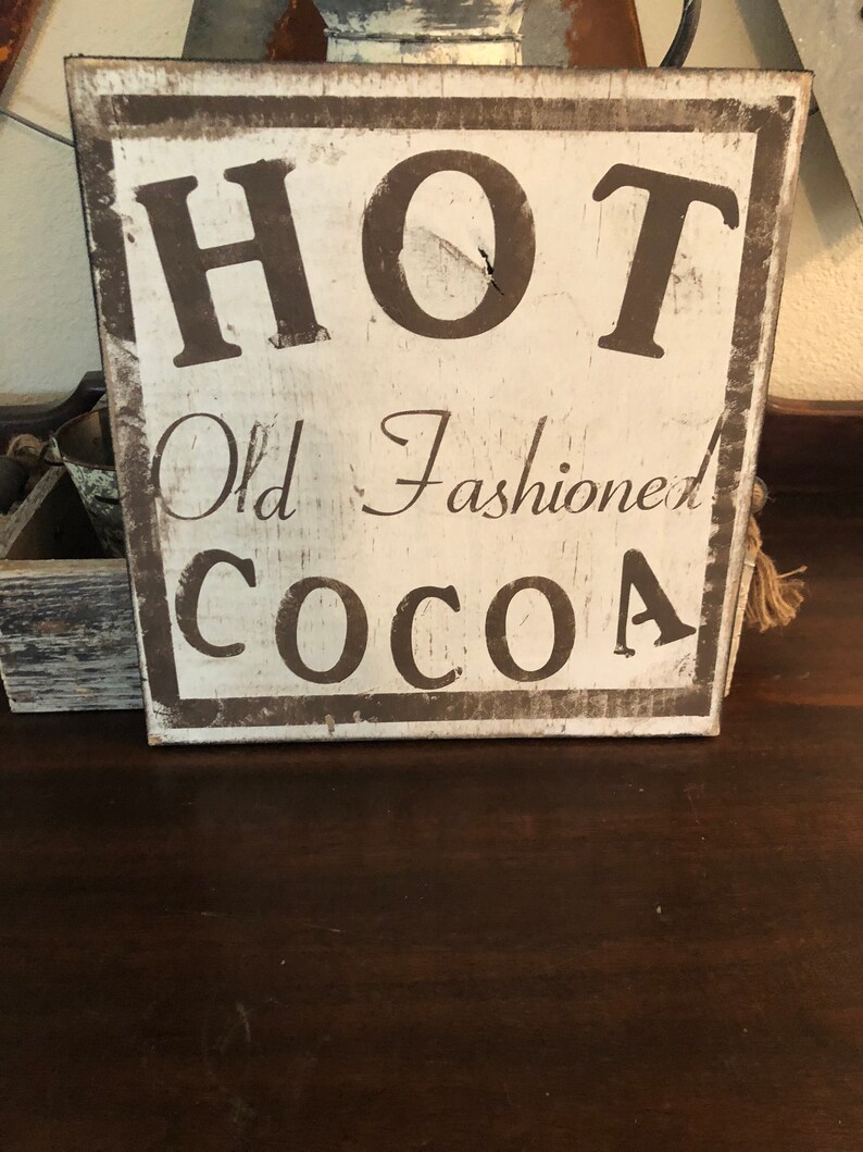 Puede incluir: Un letrero de madera r&uacute;stico con las palabras "HOT Old Fashioned COCOA" en letras marr&oacute;n oscuro. El letrero tiene un fondo blanco y un borde marr&oacute;n oscuro, con un aspecto vintage. Evoca una sensaci&oacute;n de calidez y confort.