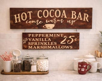 May include: Wooden signs for a hot cocoa bar, with text that reads "Hot Cocoa Bar, come warm up" and "Peppermint, Vanilla, Sprinkles, Marshmallows, 25¢". Jars of toppings and mugs are on the counter.