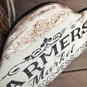 Op de afbeelding: Een wit houten bord met zwarte letters waarop "Farmers Market" en "Honey-Eggs-Pies-Butter" staat. Het bord heeft een decoratief bloemmotief in de linkerbovenhoek.