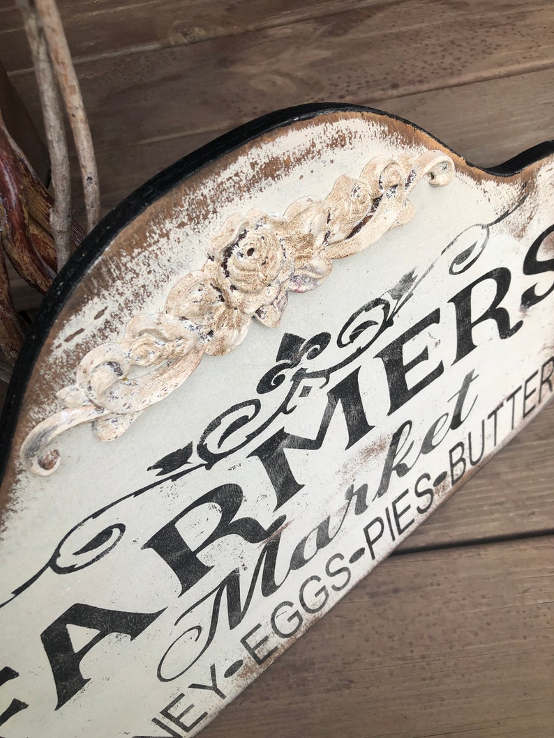 Op de afbeelding: Een wit houten bord met zwarte letters waarop "Farmers Market" en "Honey-Eggs-Pies-Butter" staat. Het bord heeft een decoratief bloemmotief in de linkerbovenhoek.