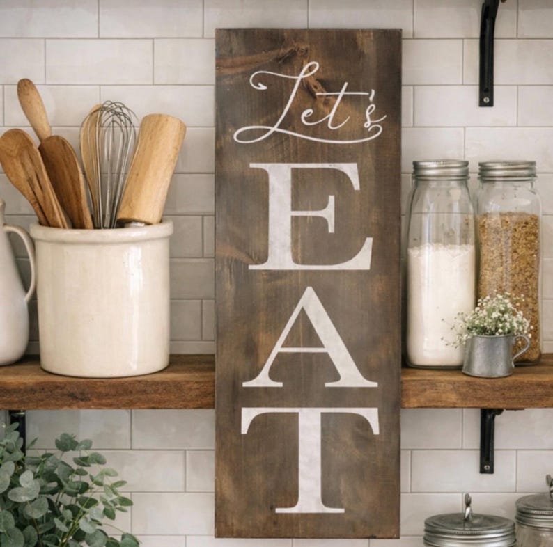 May include: A rustic wooden sign with the words "Let's EAT" in white lettering. The sign is displayed on a kitchen shelf with jars of ingredients and cooking utensils. The background features white tile.