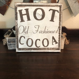 Puede incluir: Un letrero de madera r&uacute;stico con las palabras "HOT Old Fashioned COCOA" en letras marr&oacute;n oscuro. El letrero tiene un aspecto envejecido y vintage con un fondo blanco y un borde marr&oacute;n oscuro. El letrero est&aacute; colocado sobre una superficie de madera oscura.