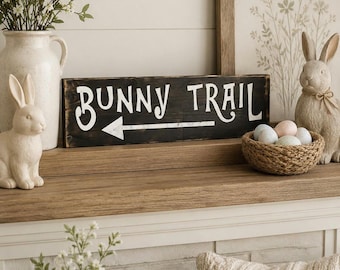 May include: A rectangular wooden sign with a dark stained finish featuring the white painted text BUNNY TRAIL and a white arrow pointing to the left. The sign is positioned horizontally, showcasing a weathered texture and clear, bold lettering.