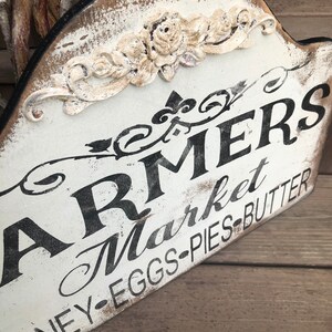 Op de afbeelding: Een wit houten bord met zwarte letters waarop "Farmers Market Honey-Eggs-Pies-Butter" staat. Het bord heeft een verweerde uitstraling en is versierd met een wit bloemmotief.
