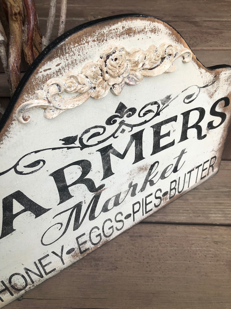 Op de afbeelding: Een wit houten bord met zwarte letters waarop "Farmers Market Honey-Eggs-Pies-Butter" staat. Het bord heeft een verweerde uitstraling en is versierd met een wit bloemmotief.