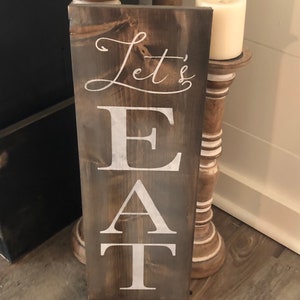 May include: A rectangular wooden sign with the words "Let's EAT" in white lettering. The sign has a dark brown stain and is leaning against a white wall. Two white candles sit on decorative wooden stands.