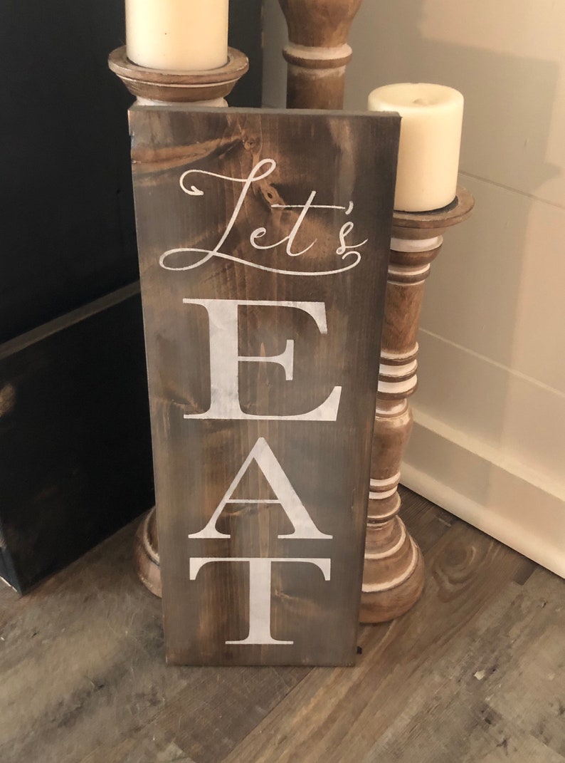 May include: A rectangular wooden sign with the words "Let's EAT" in white lettering. The sign has a dark brown stain and is leaning against a white wall. Two white candles sit on decorative wooden stands.
