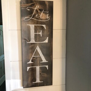 May include: A rectangular wooden sign with the words "Let's EAT" in white lettering. The sign has a dark brown stain and is mounted on a white wall. The text is in a mix of cursive and block letters.