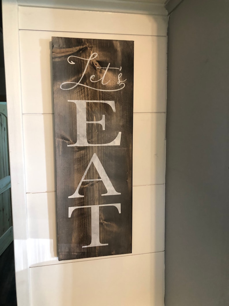 May include: A rectangular wooden sign with the words "Let's EAT" in white lettering. The sign has a dark brown stain and is mounted on a white wall. The text is in a mix of cursive and block letters.