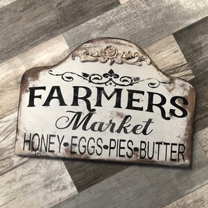 Op de afbeelding: Een wit houten bord met zwarte tekst die "Farmers Market Honey-Eggs-Pies-Butter" luidt. Het bord heeft een decoratief bloemenmotief bovenaan.