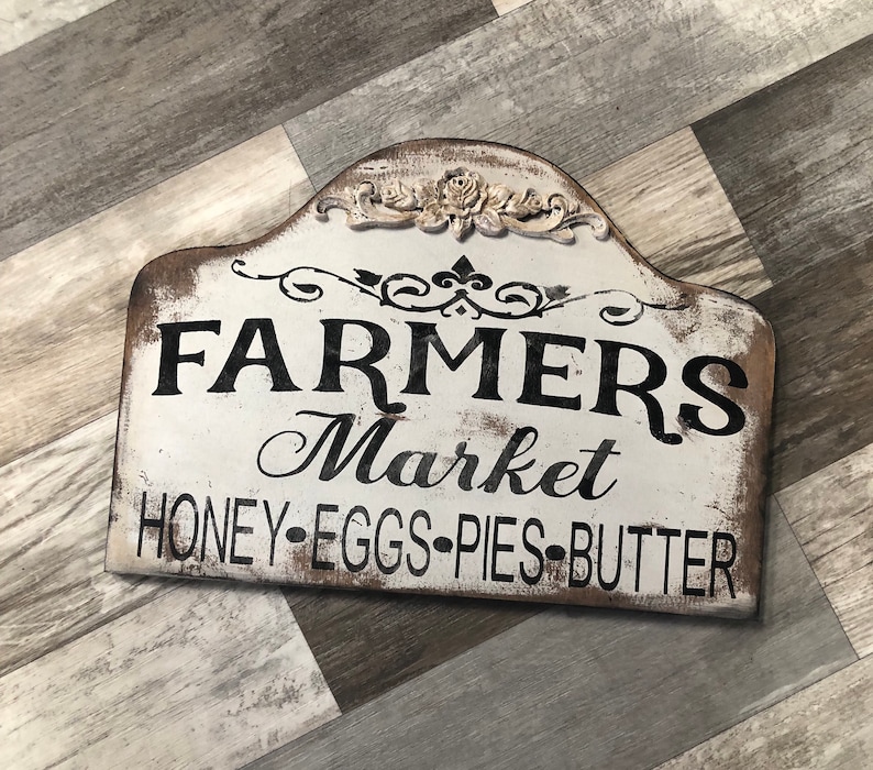 Op de afbeelding: Een wit houten bord met zwarte tekst die "Farmers Market Honey-Eggs-Pies-Butter" luidt. Het bord heeft een decoratief bloemenmotief bovenaan.