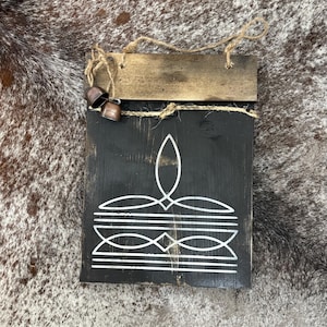 May include: A rectangular, black wooden sign with a white geometric design and a small, brown wooden top piece. Two small, copper-colored bells hang from the top. The sign is suspended by a natural twine.