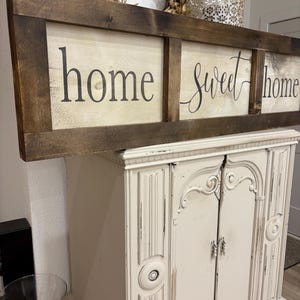 May include: A rectangular wooden sign with a dark brown stained frame and a light-colored center panel. The sign features the black text "home sweet home" divided into three sections by vertical wooden slats, with a distressed finish on the wood.