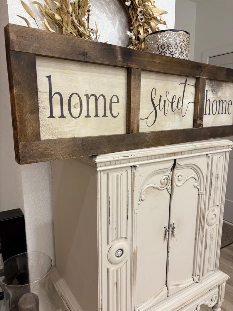May include: A rectangular wooden sign with a dark brown stained frame and a light-colored center panel. The sign features the black text "home sweet home" divided into three sections by vertical wooden slats, with a distressed finish on the wood.