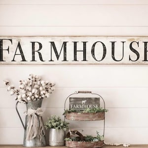 May include: A long rectangular wooden sign with a distressed white finish and black lettering that reads FARMHOUSE. The sign features vertical black lines on both ends and is displayed above a metal pitcher with cotton stems and a tiered tray.