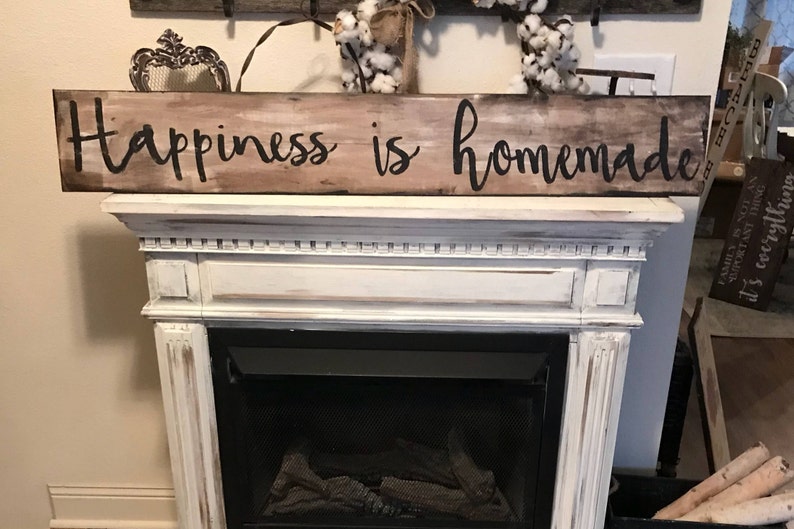 May include: A rustic wooden sign with the text "Happiness is homemade" painted in black. The sign is displayed above a white fireplace mantel.