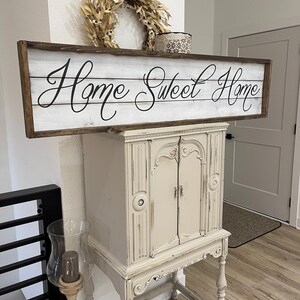 May include: A rustic wooden sign with the phrase "Home Sweet Home" in elegant script. The sign is framed in dark brown and mounted above a cream-colored cabinet. A dried wreath and decorative box sit above the sign, enhancing the cozy aesthetic.