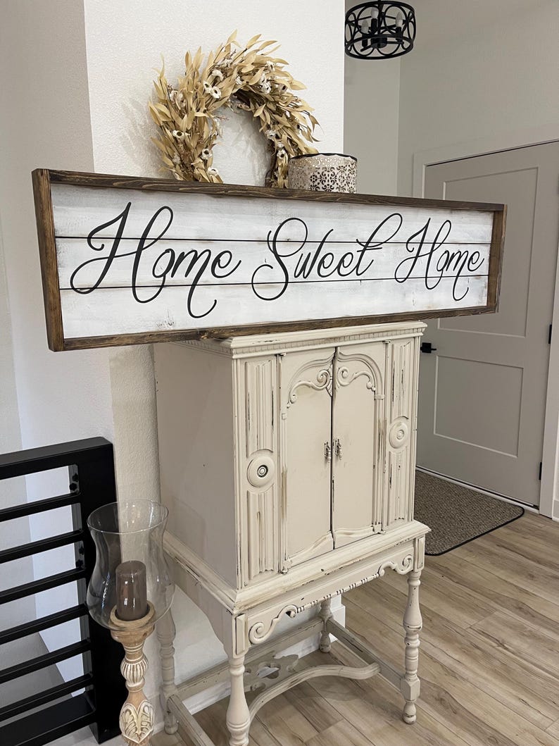 May include: A rustic wooden sign with the phrase "Home Sweet Home" in elegant script. The sign is framed in dark brown and mounted above a cream-colored cabinet. A dried wreath and decorative box sit above the sign, enhancing the cozy aesthetic.