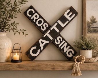 Op de afbeelding: Een decoratief X-vormig houten wandbord met een verweerde zwarte afwerking en witte letters met de tekst CROS en CATTLE SING. De twee kruisende planken vormen een kruisontwerp en vertonen over het gehele oppervlak een verweerde textuur.