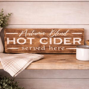 May include: A rustic wooden sign with the text "Autumn Blend HOT CIDER served here" sits on a wooden shelf. A white pitcher, a plaid napkin, and a white mug are also on the shelf, along with a basket of red apples.