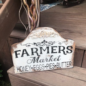 Op de afbeelding: Een wit houten bord met zwarte letters waarop staat "Farmers Market Honey-Eggs-Pies-Butter". Het bord heeft een verweerde uitstraling en is versierd met een wit bloemmotief.