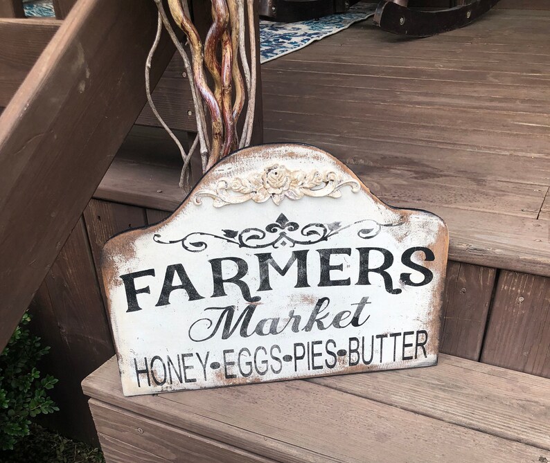Op de afbeelding: Een wit houten bord met zwarte letters waarop staat "Farmers Market Honey-Eggs-Pies-Butter". Het bord heeft een verweerde uitstraling en is versierd met een wit bloemmotief.