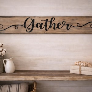 May include: A wooden sign with the word "Gather" in black script lettering hangs above a shelf. The shelf displays a galvanized metal bucket with cotton stems, a white pitcher, books, and a lantern with a lit candle.
