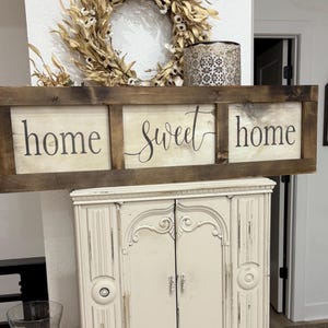 May include: A rectangular wooden sign with a dark brown stained frame and three white panels. The text "home sweet home" is printed in black lettering across the panels, with the word "sweet" in a cursive script and "home" in a serif font.