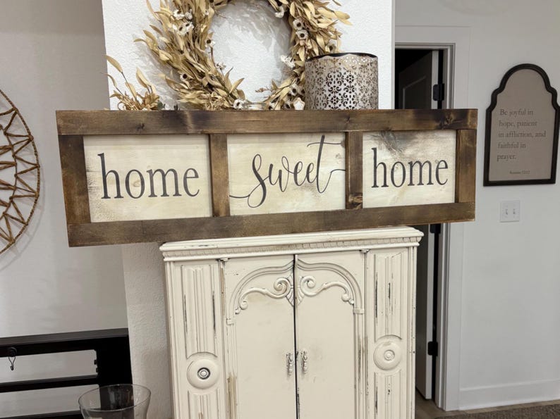 May include: A rectangular wooden sign with a dark brown stained frame and three white panels. The text "home sweet home" is printed in black lettering across the panels, with the word "sweet" in a cursive script and "home" in a serif font.