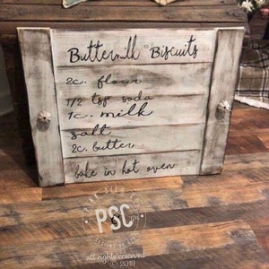Może przedstawiać: Biały drewniany znak z czarnym napisem "Buttermilk Biscuits" i przepisem na ciasteczka z maślanki. Znak jest postarzony i ma rustykalny wygląd.