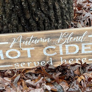 May include: A rectangular wooden sign with the words "Autumn Blend HOT CIDER served here" in white lettering. The sign is set against a backdrop of fallen leaves and a tree trunk.