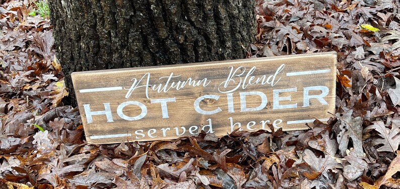 May include: A rectangular wooden sign with the words "Autumn Blend HOT CIDER served here" in white lettering. The sign is set against a backdrop of fallen leaves and a tree trunk.