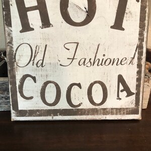 Puede incluir: Un letrero rectangular de madera envejecida con las palabras "HOT Old Fashioned COCOA" en letras marr&oacute;n oscuro. El letrero tiene un fondo blanco con un borde marr&oacute;n oscuro, evocando una est&eacute;tica vintage.
