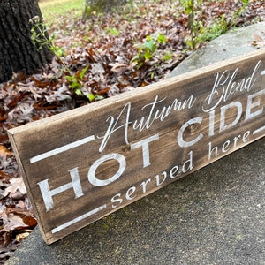 May include: A rectangular wooden sign with the words "Autumn Blend HOT CIDER served here" in white lettering. The sign has a rustic, weathered appearance with a dark brown stain.
