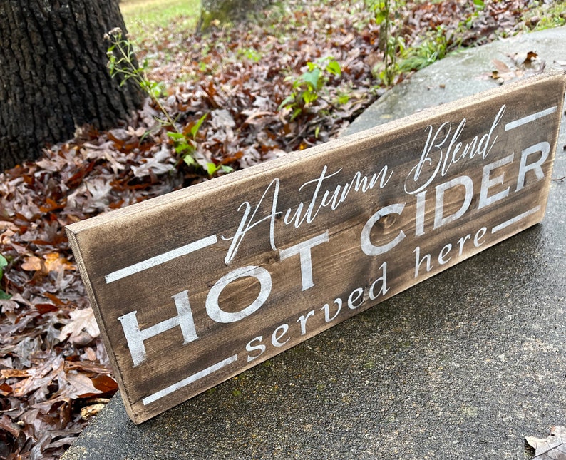 May include: A rectangular wooden sign with the words "Autumn Blend HOT CIDER served here" in white lettering. The sign has a rustic, weathered appearance with a dark brown stain.